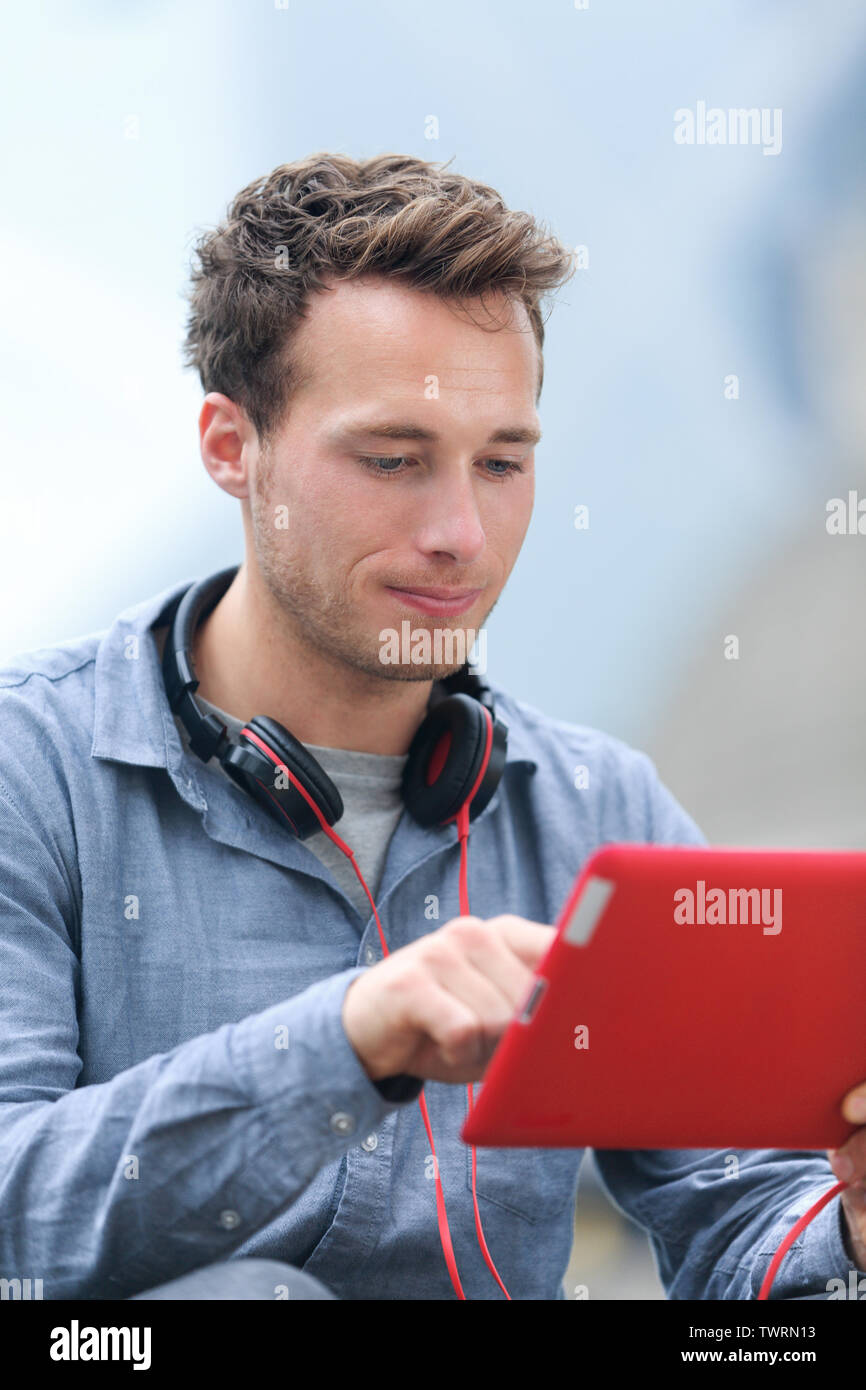 Urban young professional man using tablet computer sitting outside using app on 4g wireless device wearing headphones. Casual young urban professional male in his late 20s. Stock Photo