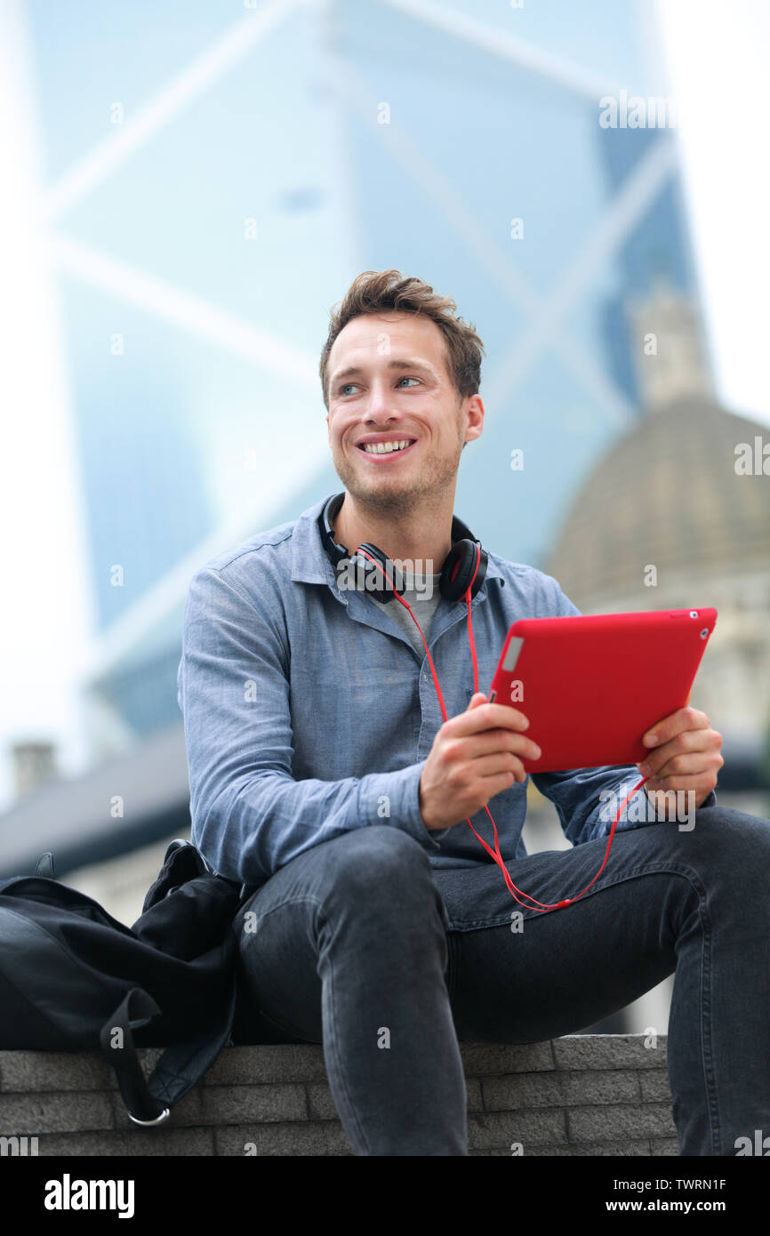 Urban young professional man using tablet computer sitting in Hong Kong outside using app on 4g wireless device wearing headphones. Casual young urban professional male in his late 20s. Stock Photo