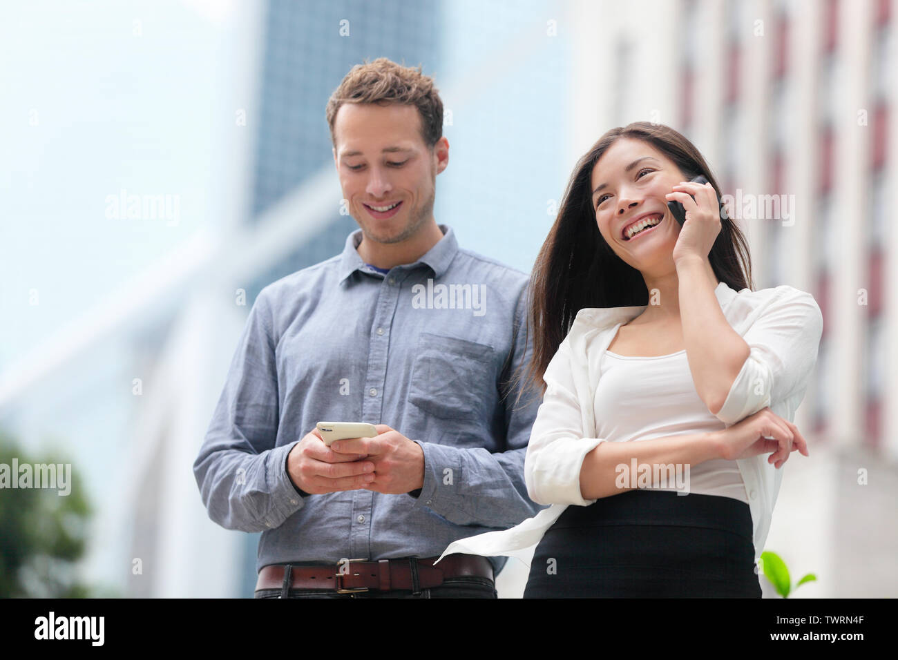Young urban professionals business people on smartphones in Hong Kong. Businessman using app on smartphone and businesswoman talking having conversation on smart phone in Hong Kong Central. Stock Photo