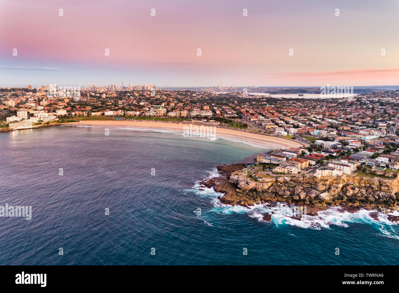 North Bondi sandstone cliff of headland guarding famours Bondi Beach of Sydney from open waves of Pacific ocean in aerial sunrise view towards distant Stock Photo