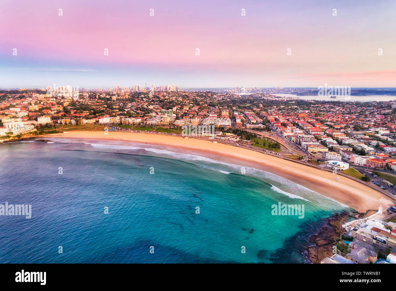Clean arc of wide sandy Bondi Beach in Sydney at sunrise with pink sky and calm smooth waves floating surfers and surrounded by Eastern suburbs. Stock Photo