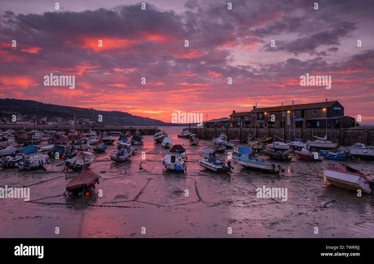 Lyme Regis, Dorset, UK. 23rd June 2019. UK Weather: The sky over the historic Cobb harbour glows with vibrant red sunrise colours as firey clouds bring a break in the sunshine at Lyme Regis on Sunday morning. Credit: Celia McMahon/Alamy Live News. Stock Photo