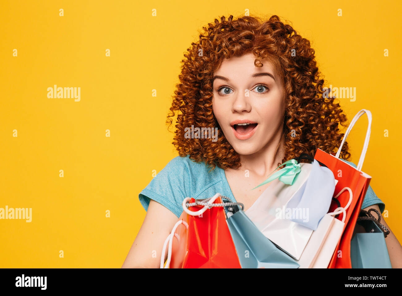 amazing Sale. Curly haired woman with shocked face holding bags with purchase on yellow background Stock Photo