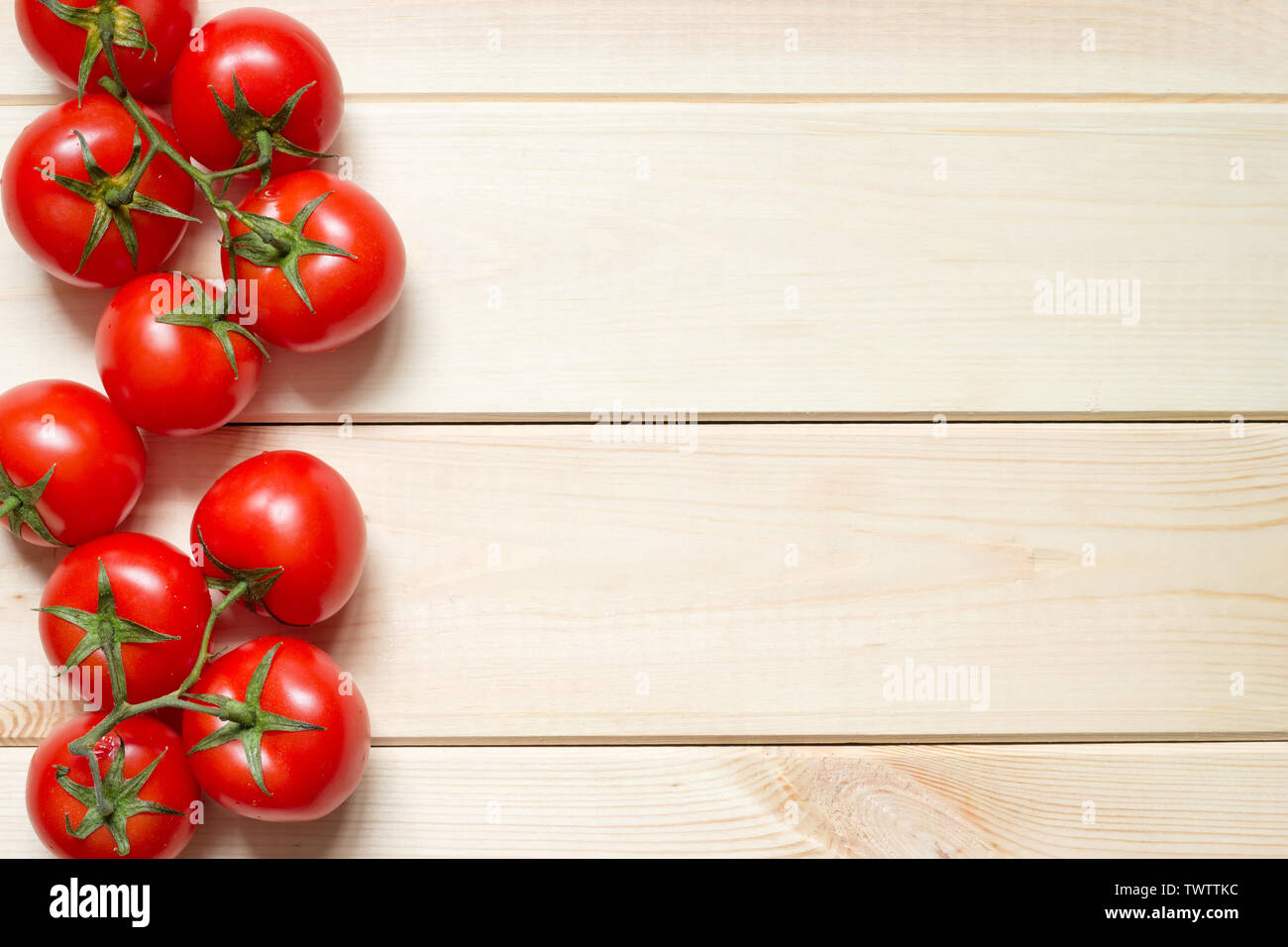 Red ripened tomatoes laid out at left side on light wooden table with copy space. Many washed vegetables on green branch with water drops on it. Top v Stock Photo