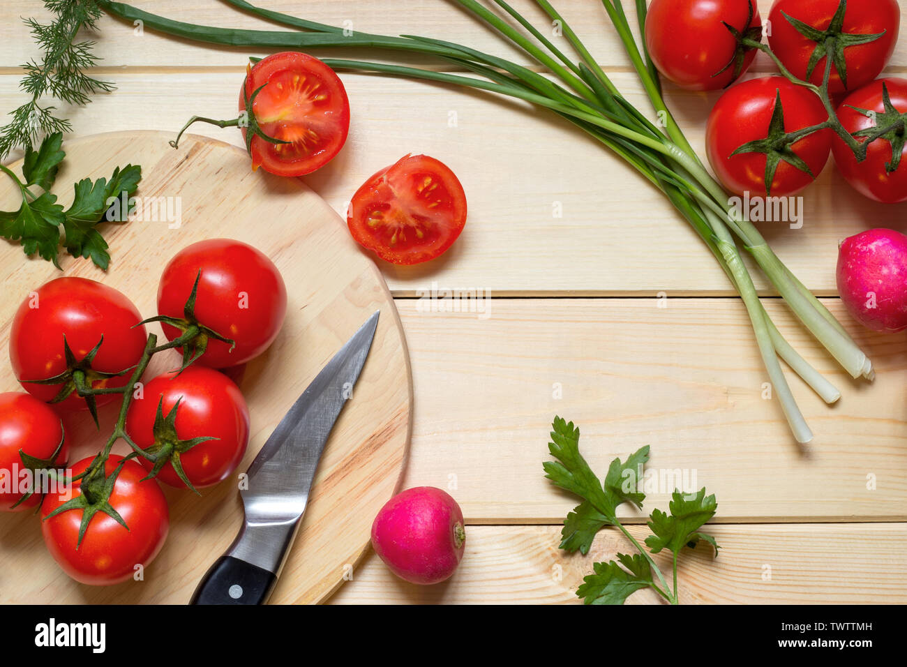 Healthy food cooking concept. Fresh tomatoes on branch, radish, onion, dill and parsley laid out on rustic light wooden table. Knife on kitchen board. Stock Photo