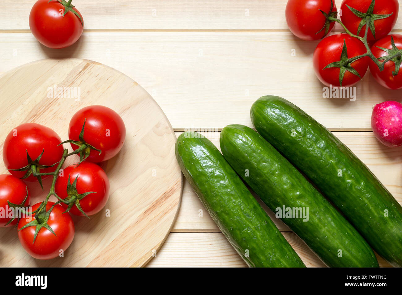 Fresh organic food ingredients on kitchen wooden table. Red cherry tomatoes, green cucumbers and radish. Round kitchen board. Fresh vegetables. Raw fo Stock Photo