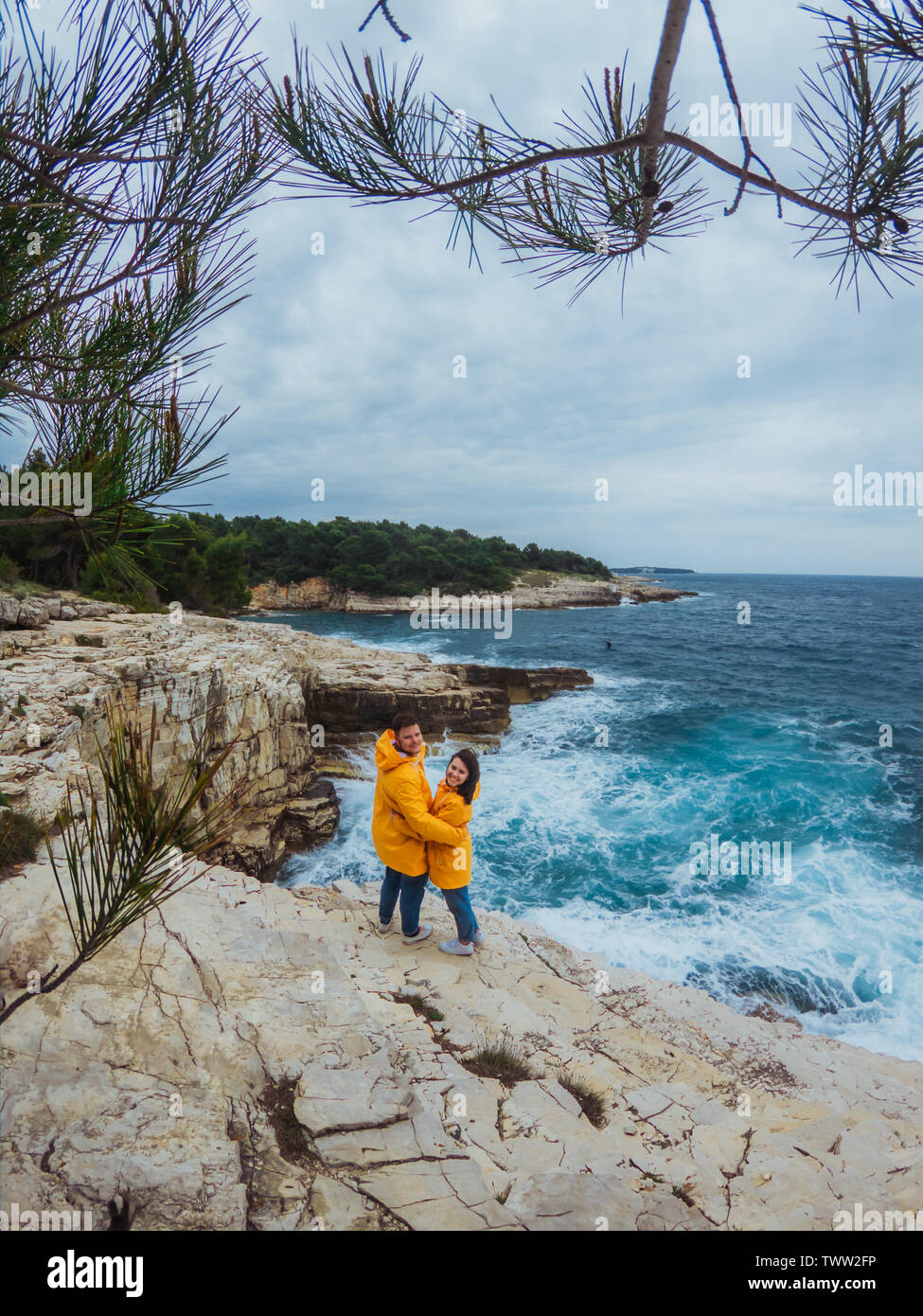 smiling couple in yellow raincoats at the edge of the cliff looking at ...