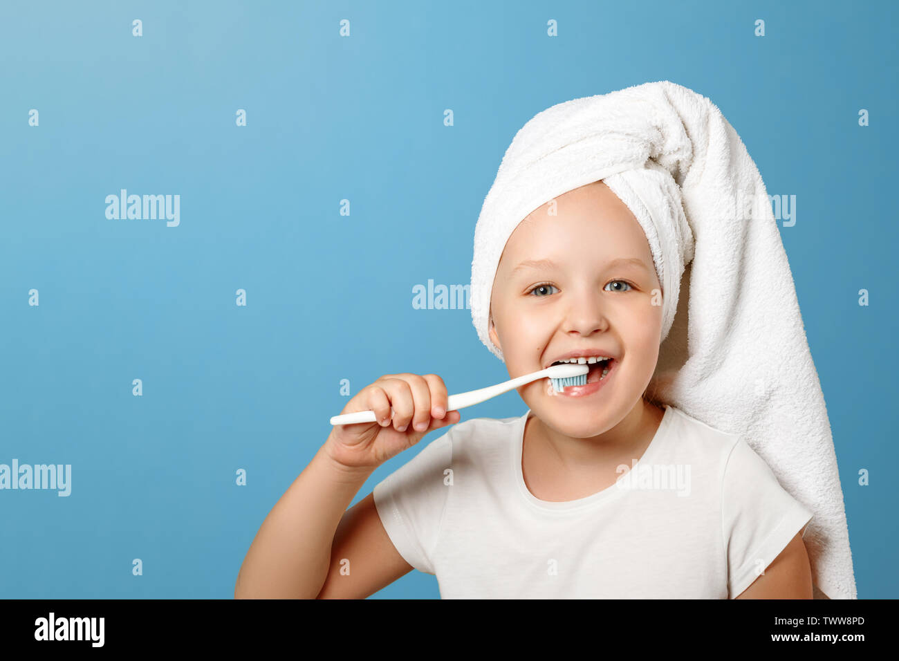 Closeup portrait of a little girl on a blue background. A child with a white towel on his head brushing his teeth. The concept of daily hygiene. Stock Photo