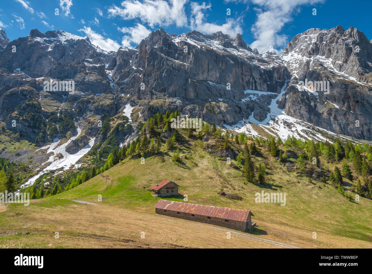 Imposing mountain range towers above alpine meadows and mountain hut lower in the valley. Avalanche prone slopes in the Dolomites. Lush pasture. Stock Photo