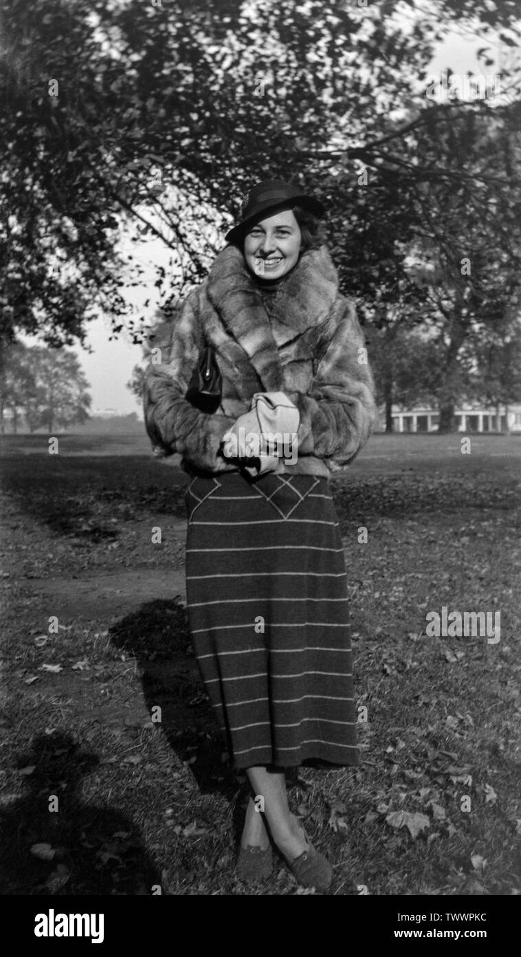 A vintage black and white photograph taken during the 1930s in England, showing a woman, posing to a camera, outside in a park, wearing a fur jacket, and typical fashion of the period. Stock Photo