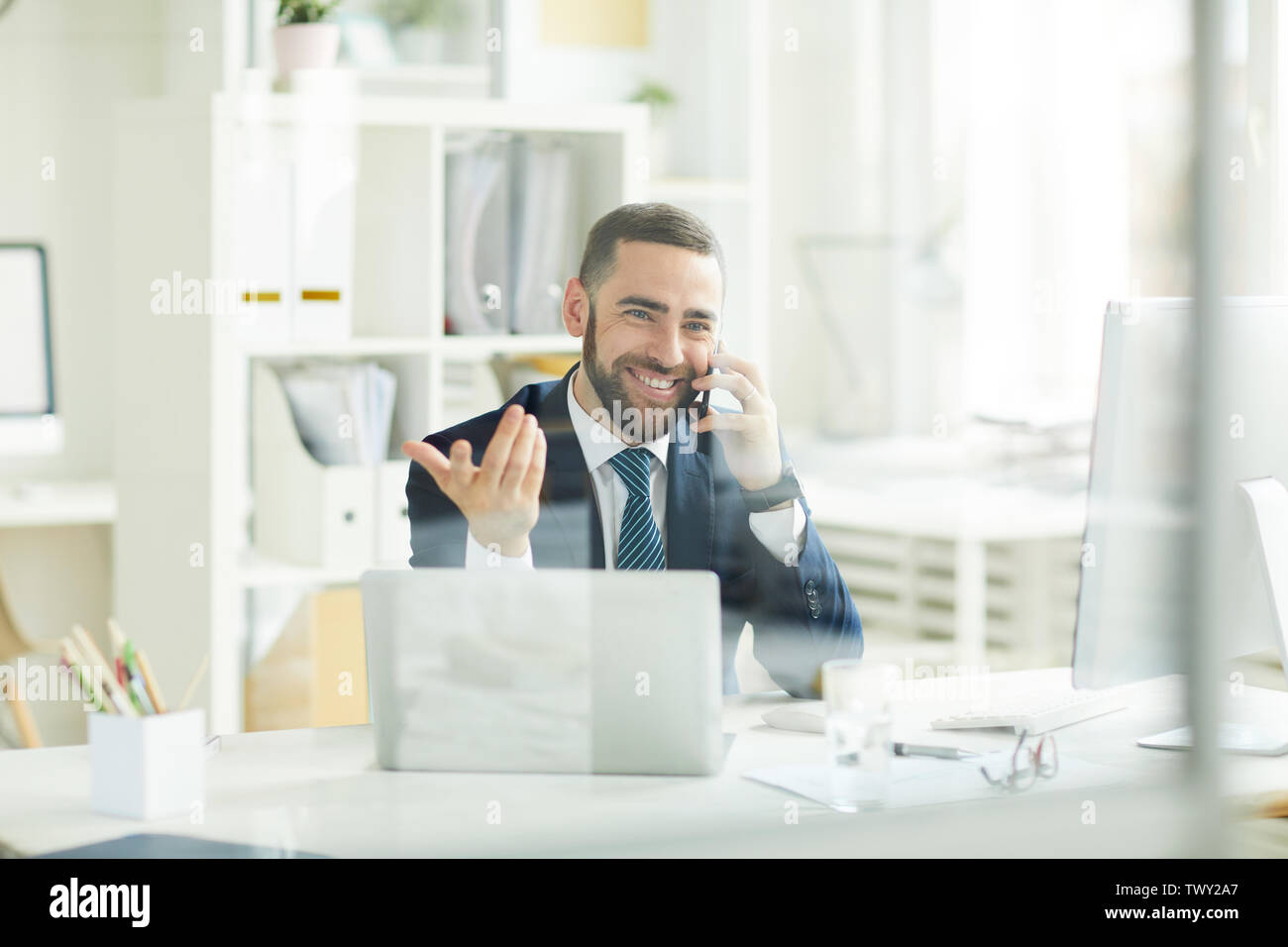 Positive confident young bearded trader in formal suit sitting at table and gesturing hand while consoling customer about financial assets on phone Stock Photo