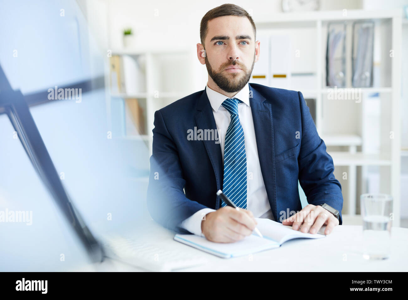 Serious purposeful young bearded businessman in formal suit sitting at table and making notes in diary while planning schedule Stock Photo