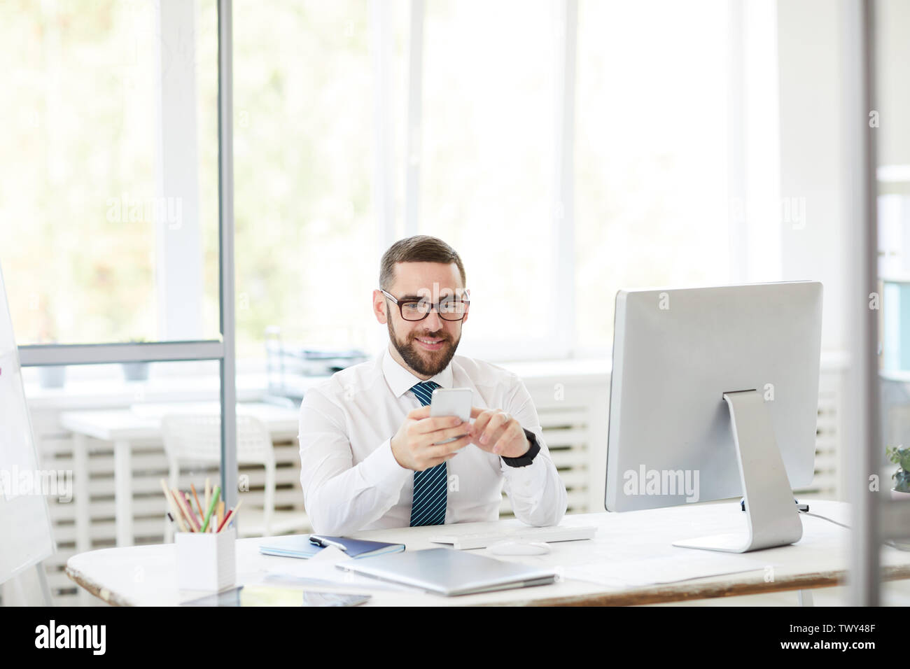 Smiling confident handsome young bearded manager in eyeglasses sitting at table and using smartphone while working in office Stock Photo