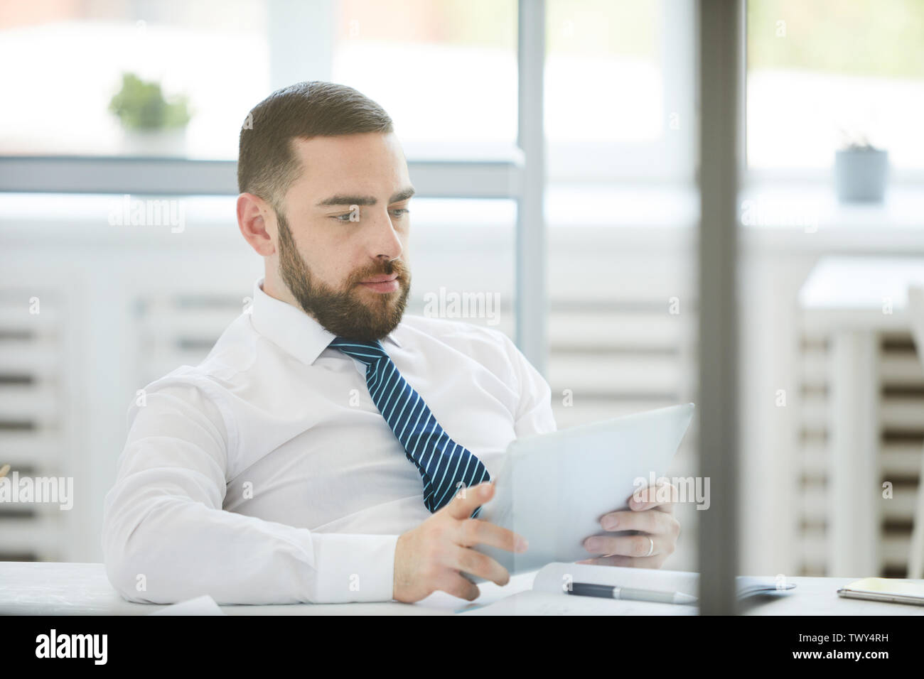 Serious busy young bearded manager sitting in relaxed pose at table in office and using tablet while checking data Stock Photo