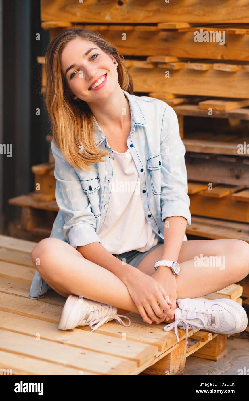 Pretty smiling girl, wearing jeans, shorts and shirt posing on wood pallet background during a sunny day. Stock Photo