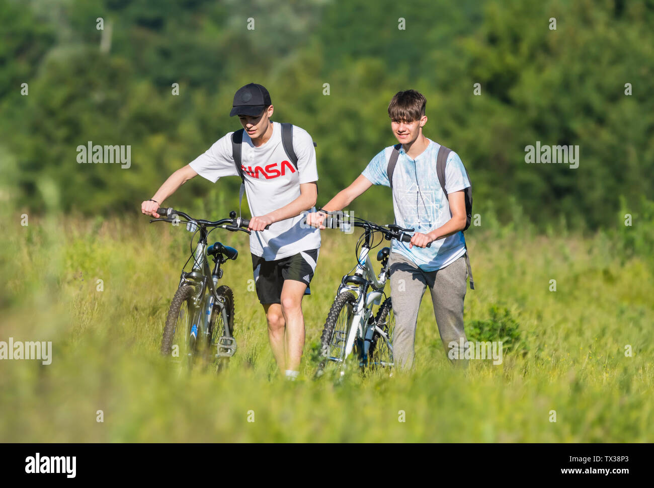 Pair of teenage friends pushing bicycles uphill through grass in the countryside in West Sussex, England, UK. Stock Photo
