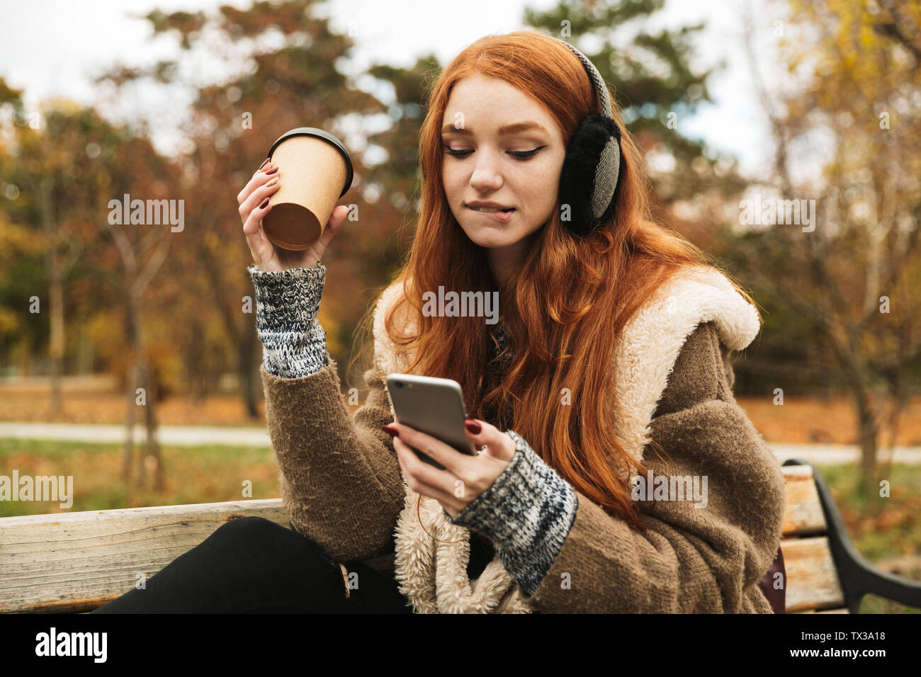 Lovely redheaded young girl listening to music with headphones while sitting on a bench, using mobile phone Stock Photo