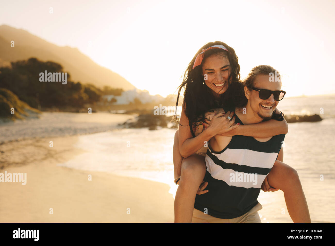 Young man and woman on vacation on a beautiful summer's day. Happy man carrying a woman on his back walking on beach. Stock Photo