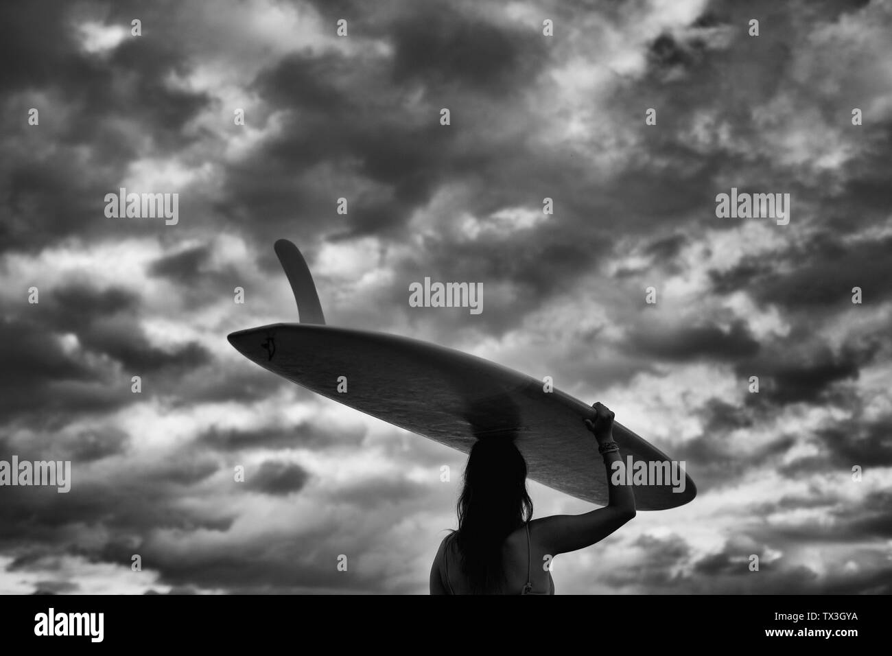 Female surfer carrying long board overhead under cloudy sky Stock Photo