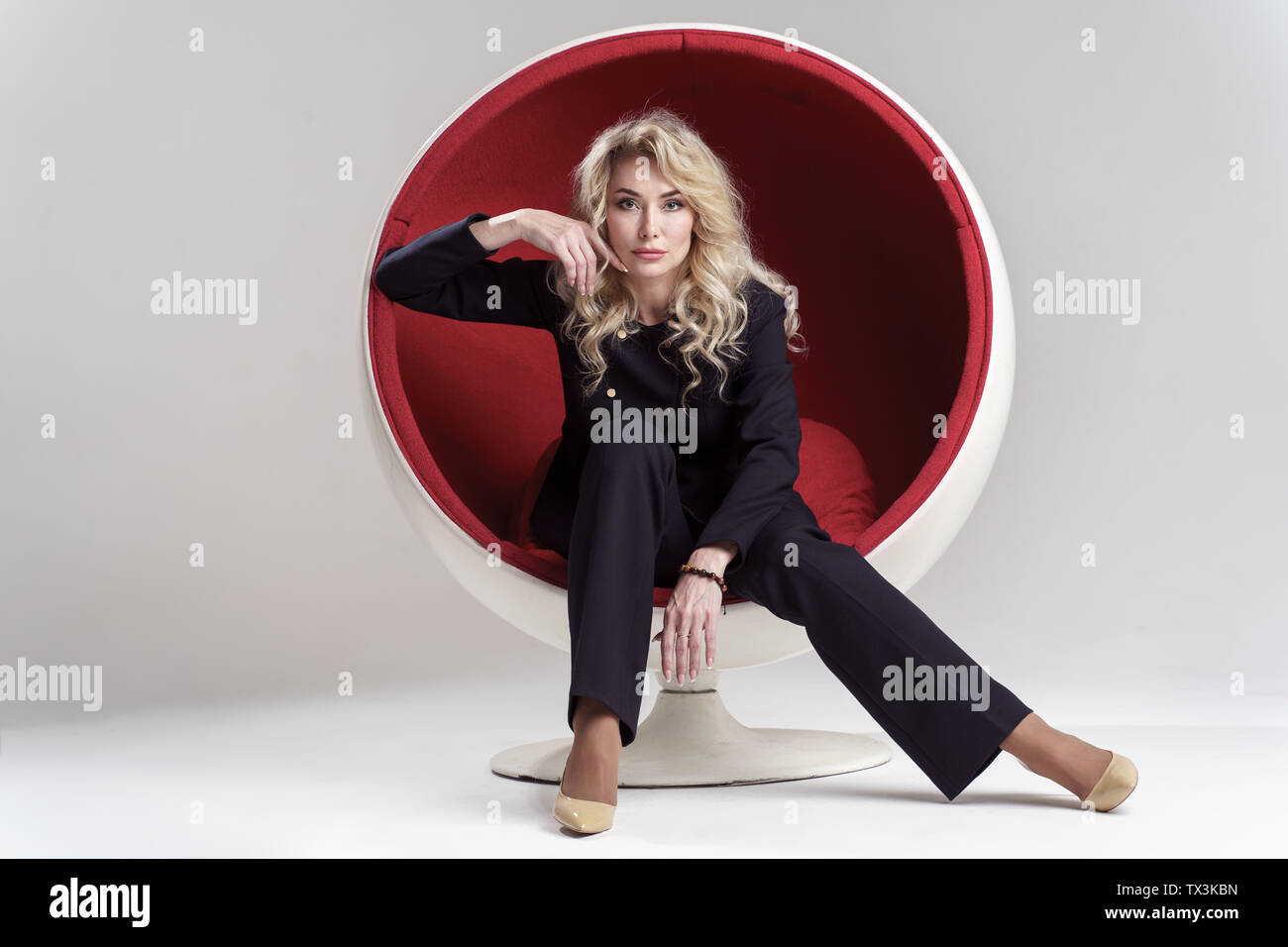 Image of blonde woman with long curly hair in black suit and beige shoes looking in camera while sitting in round chair isolated on white background Stock Photo