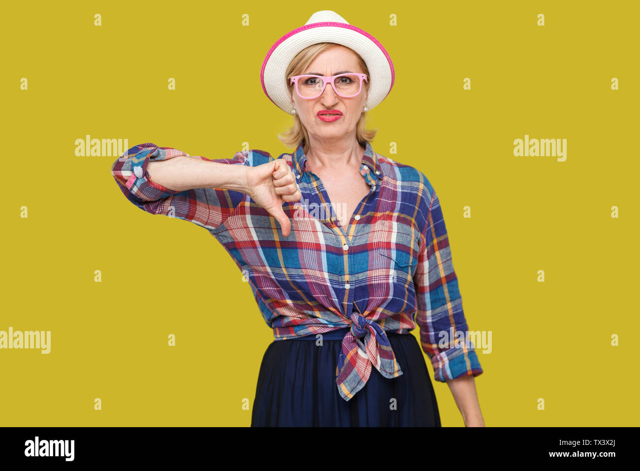 Dislike. Portrait of sad dissatisfied modern stylish mature woman in casual style with hat and eyeglasses standing looking at camera with thumb down. Stock Photo