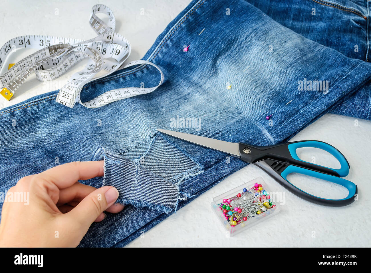 Woman hand shows a large hole on pant leg of a blue jeans folded in half.  Jeans are prepared for cutting out for making denim shorts. With scissors,  p Stock Photo -