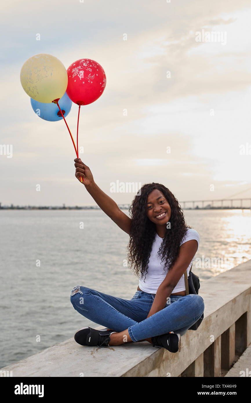 Mocambique, Maputo, portrait of happy young woman with three balloons sitting in front of the sea Stock Photo