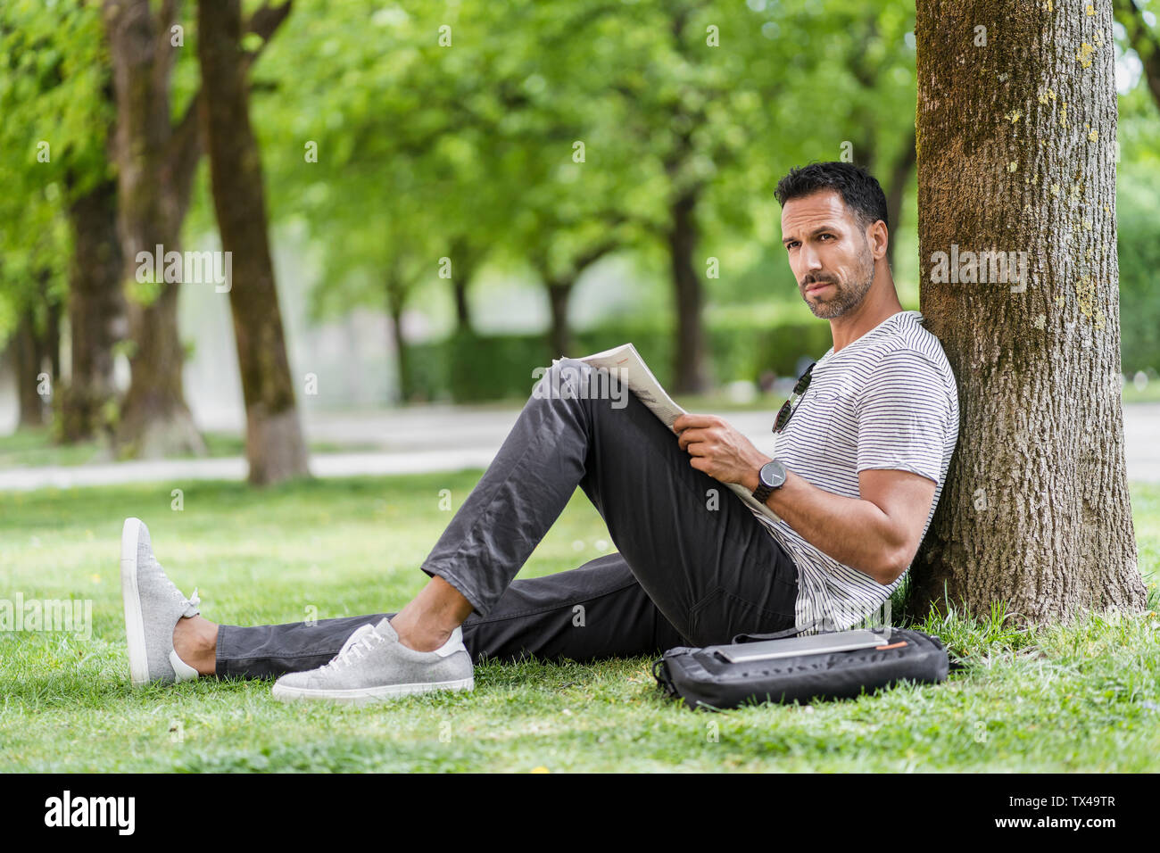 Man leaning against a tree in park reading newspaper Stock Photo