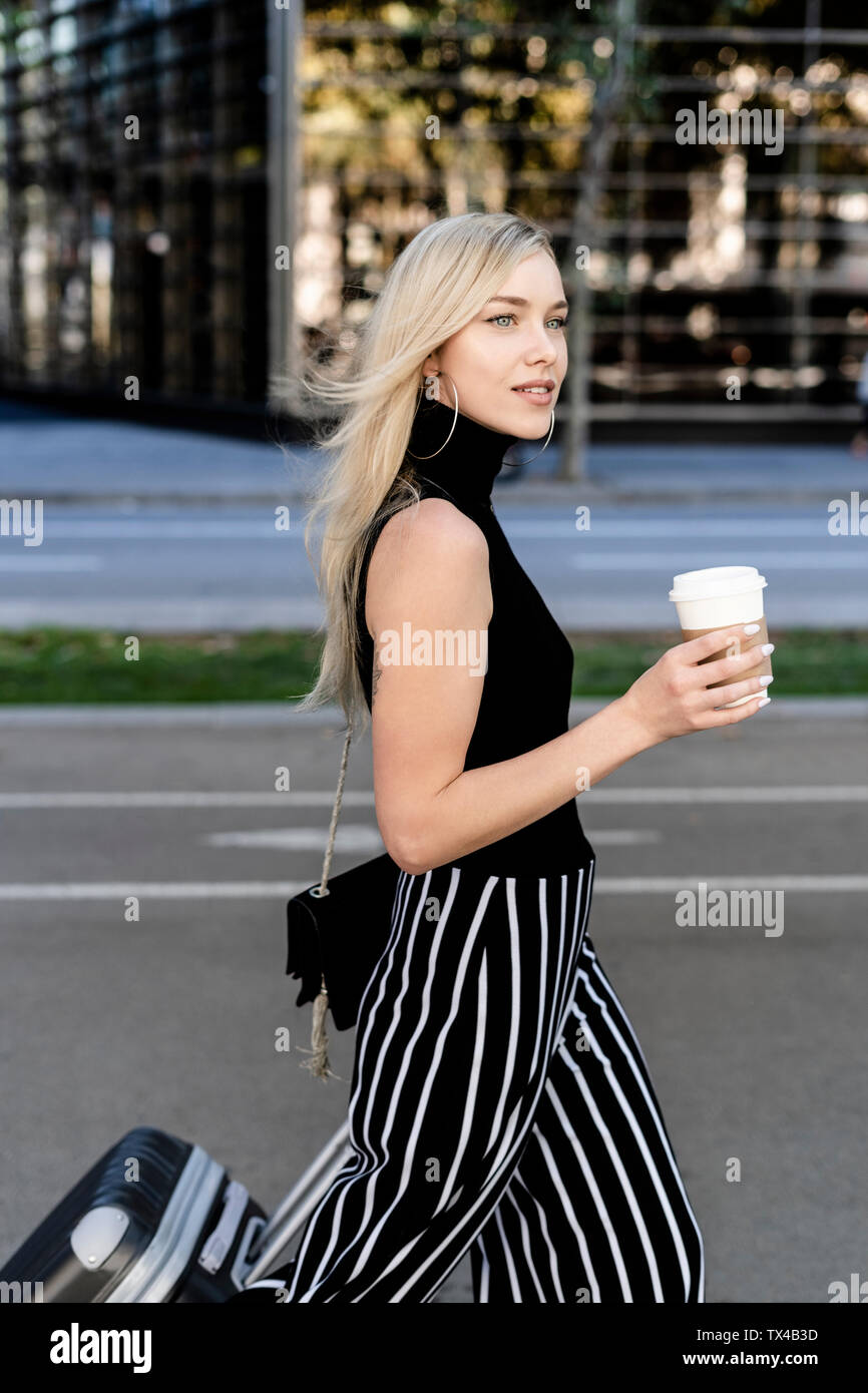 Portrait of smiling blond woman with coffee to go and wheeled luggage Stock Photo