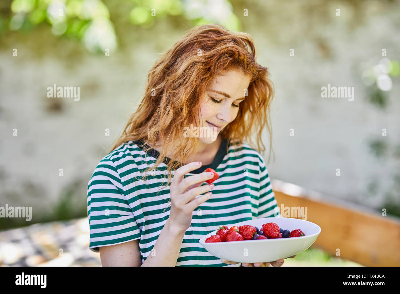 Portrait of smiling redheaded young woman with bowl of berries in the garden Stock Photo