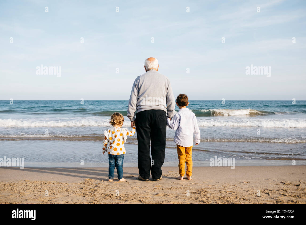 Back view of grandfather standing hand in hand on the beach with his grandchildren watching the sea Stock Photo