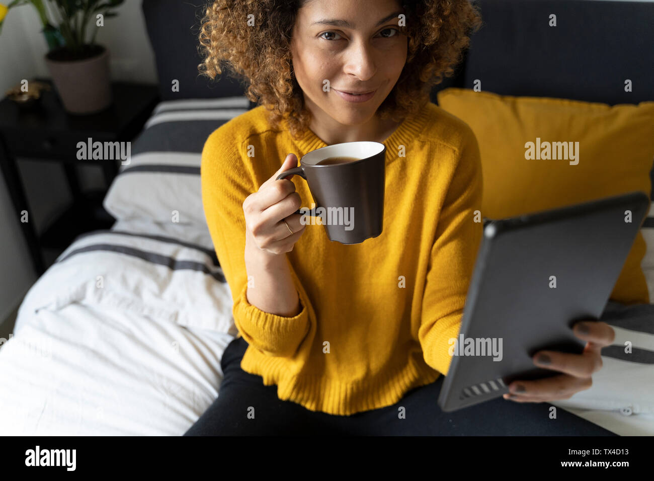 Woman sitting on bed, taking a break, drinking coffee, treading on digital tablet Stock Photo