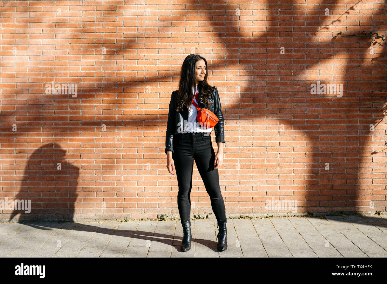 Young woman with a red bag waiting at a brick wall in the background and a shadow of tree Stock Photo