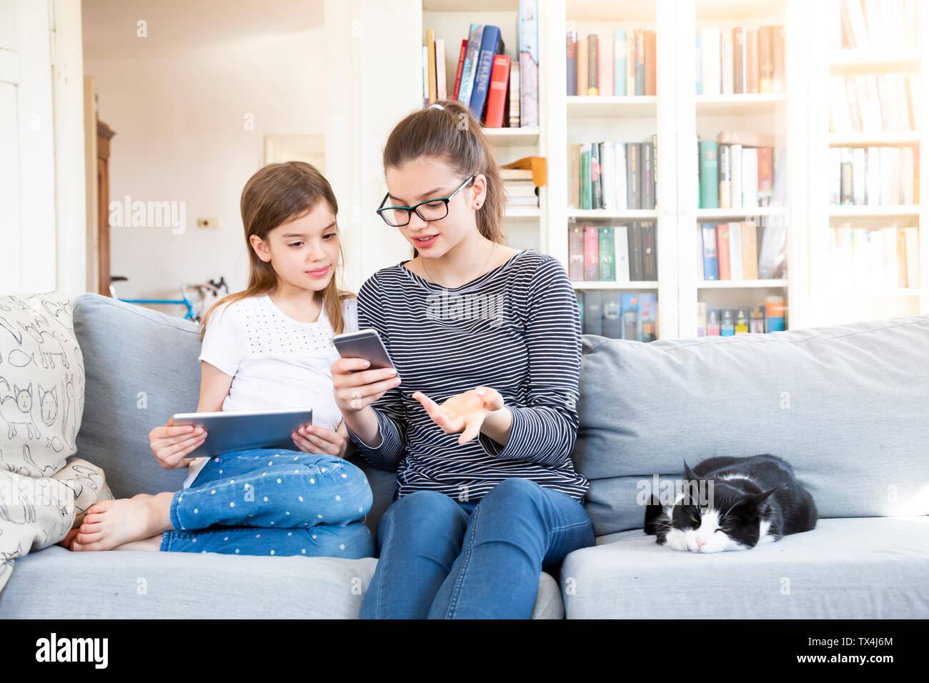 Two sisters sitting on the couch at home using electronic devices Stock Photo