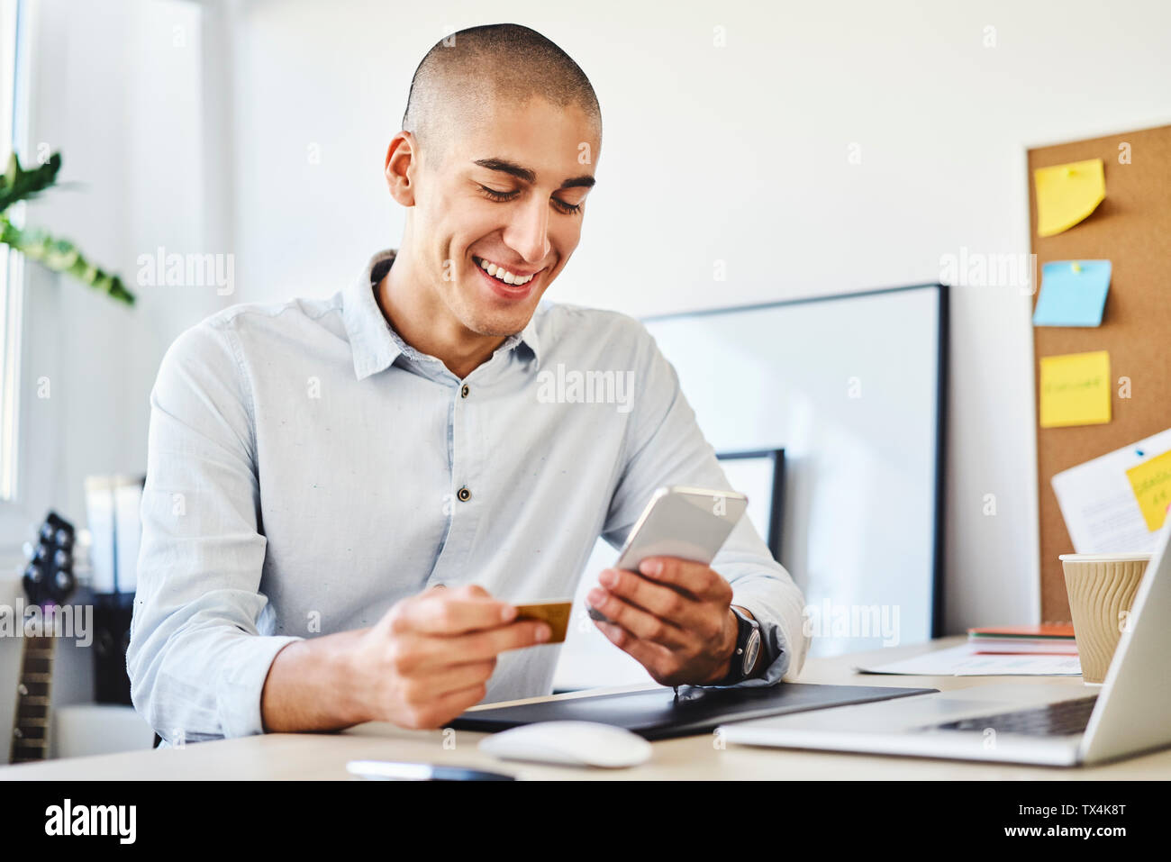 Young man sitting in home office paying online with credit card and smartphone Stock Photo