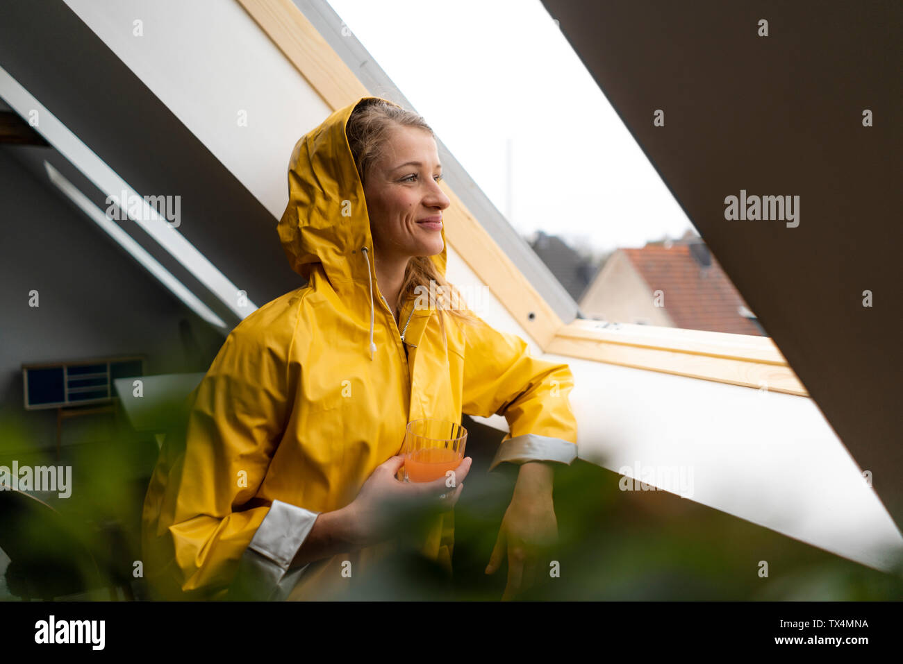 Smiling young woman in raincoat looking out of attic window Stock Photo