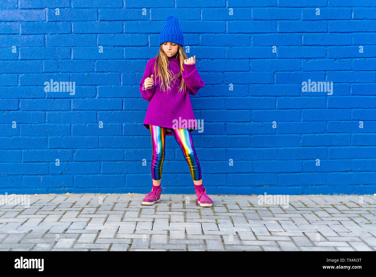 Girl wearing blue cap and oversized pink pullover standing in front of blue wall dancing Stock Photo