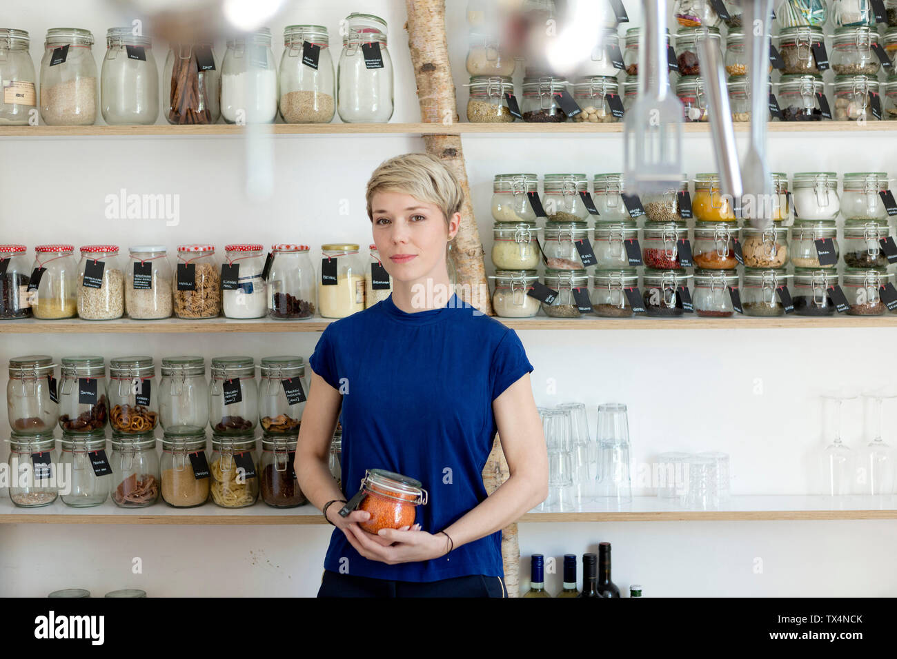 Portrait of woman holding jar in front of spice shelf in kitchen Stock Photo
