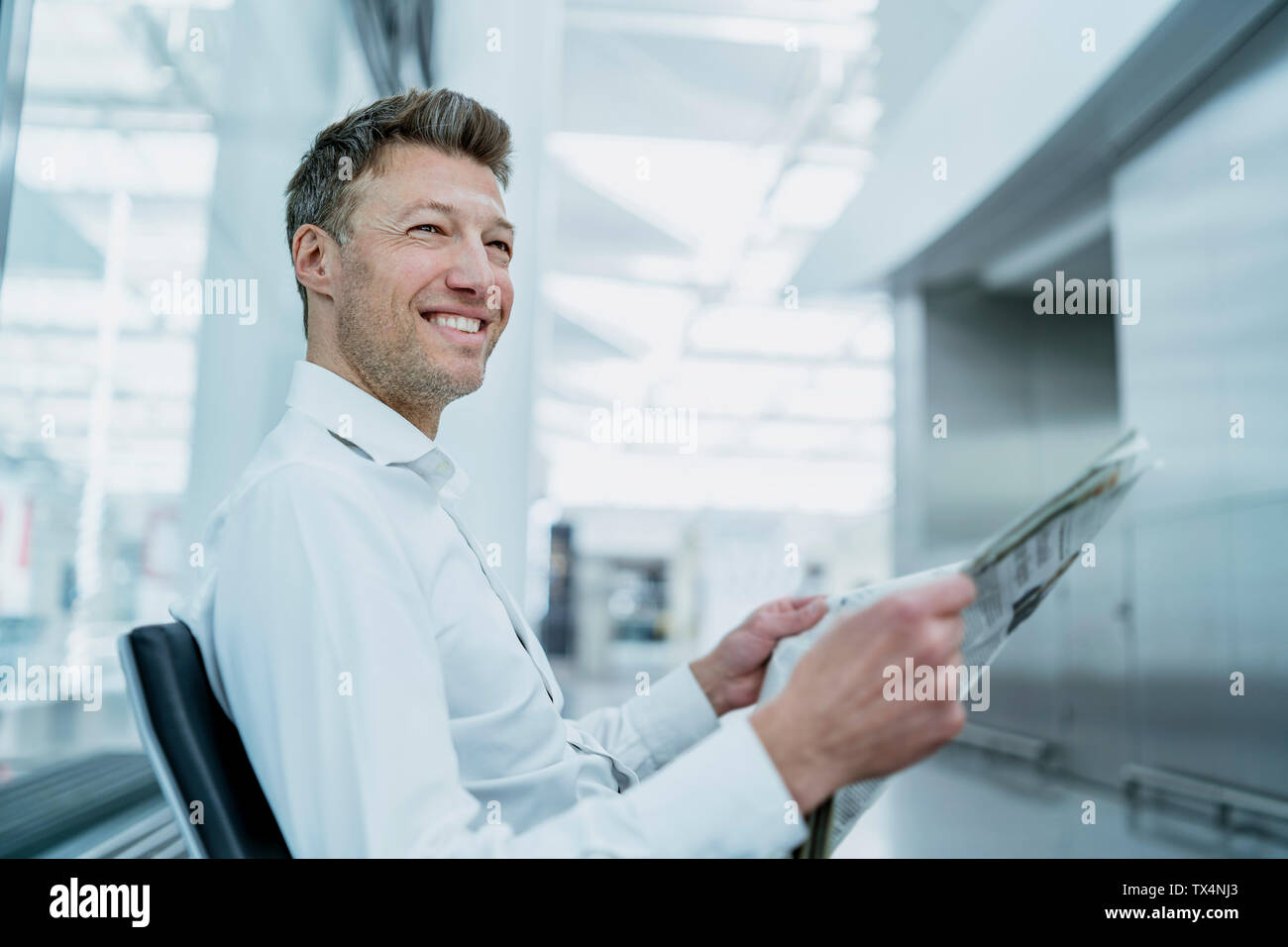 Smiling businessman sitting in waiting area with newspaper Stock Photo