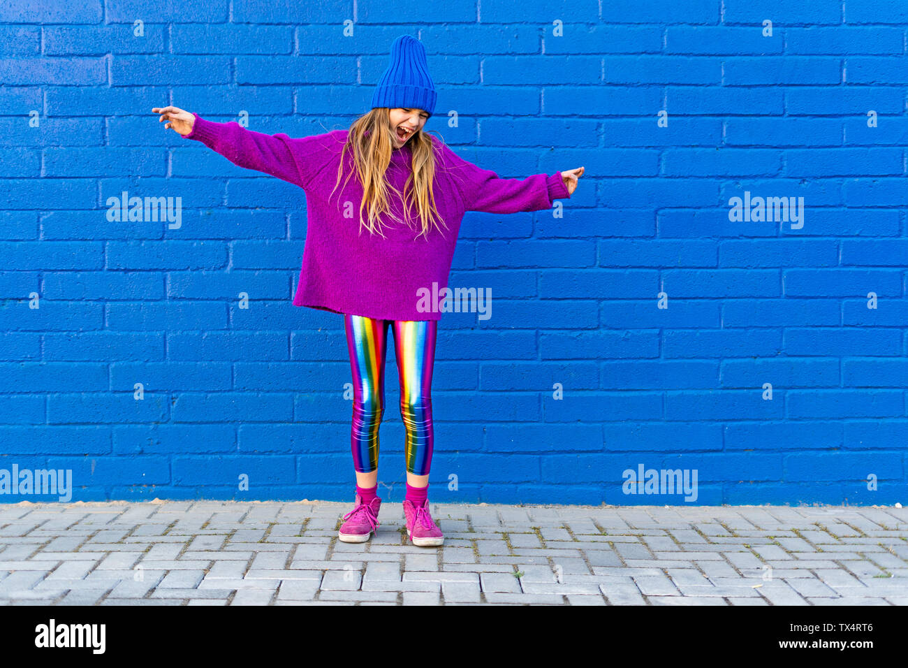 Girl wearing blue cap and oversized pink pullover standing in front of blue wall singing Stock Photo