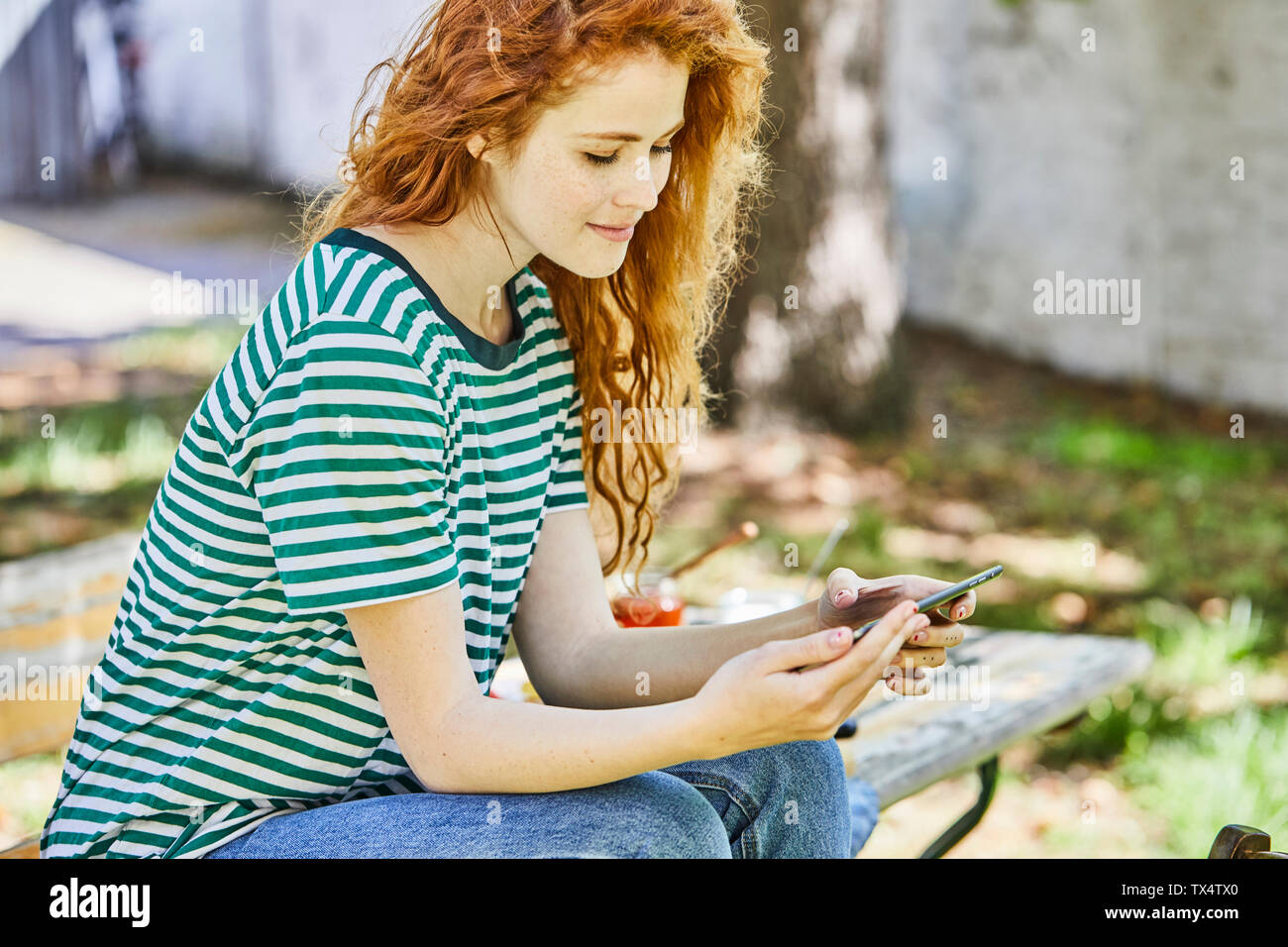 Smiling redheaded young woman  sitting on bench in the garden looking at cell phone Stock Photo