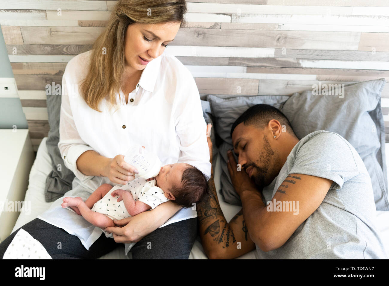 Mother bottlefeeding her newborn baby in bed with father sleeping