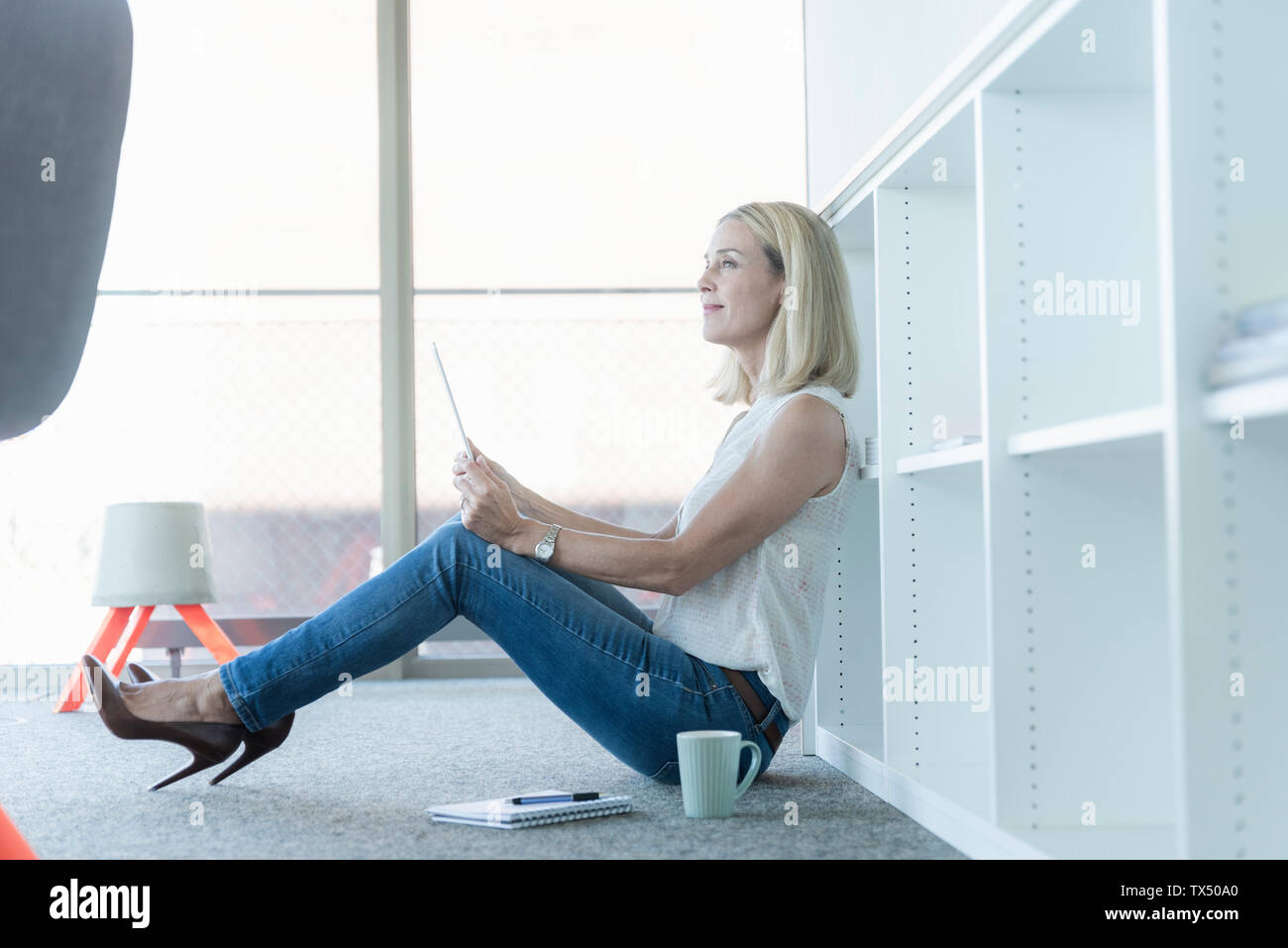 Casual businesswoman sitting on the floor in office using tablet Stock Photo