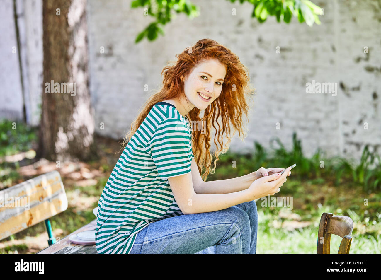 Portrait of smiling redheaded young woman with smartphone sitting on bench in the garden Stock Photo