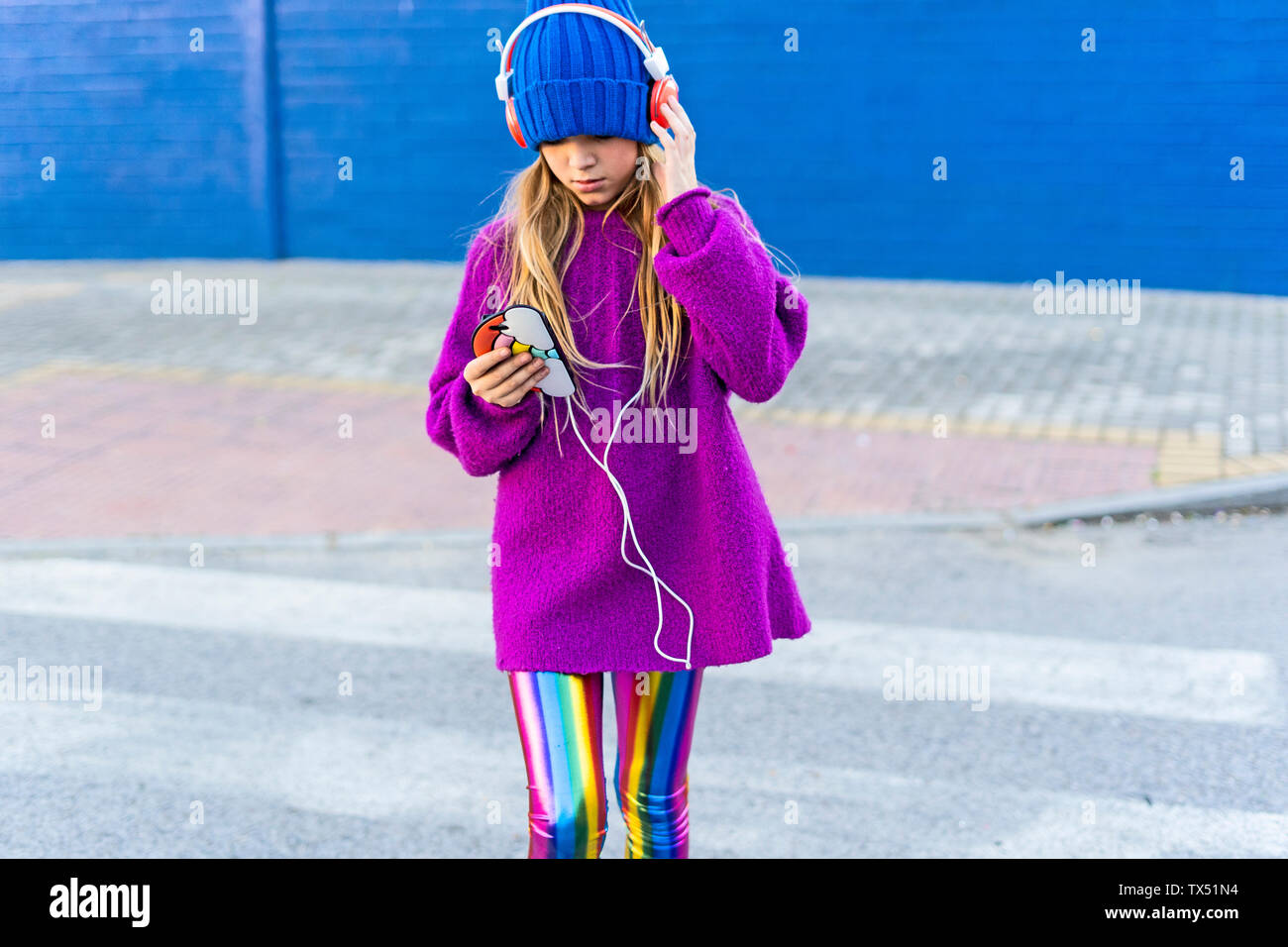 Girl wearing blue cap and oversized pink pullover listening music with headphones and smartphone Stock Photo