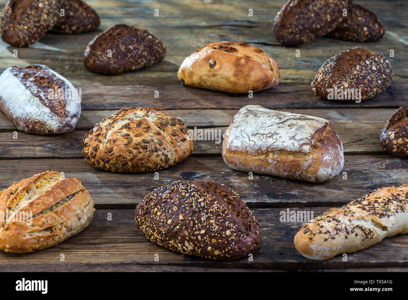 Several small multi grain different shaped bread sprinkled with whole sunflower seeds, flax and sesame seeds and wheat and barley spikes on old brown wooden table Stock Photo