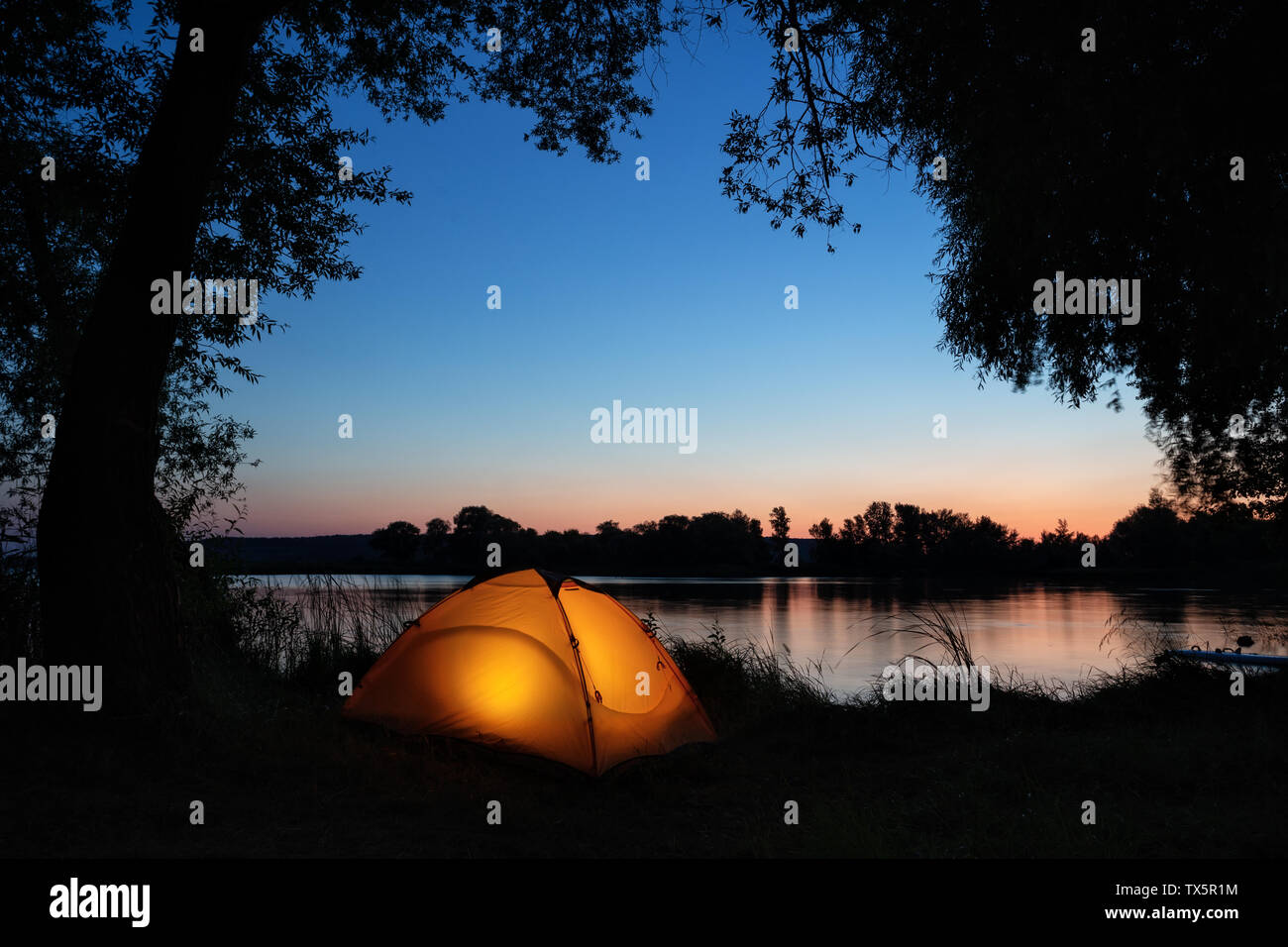 Illuminated from inside orange tent on the shore of lake among silhouettes of trees Stock Photo