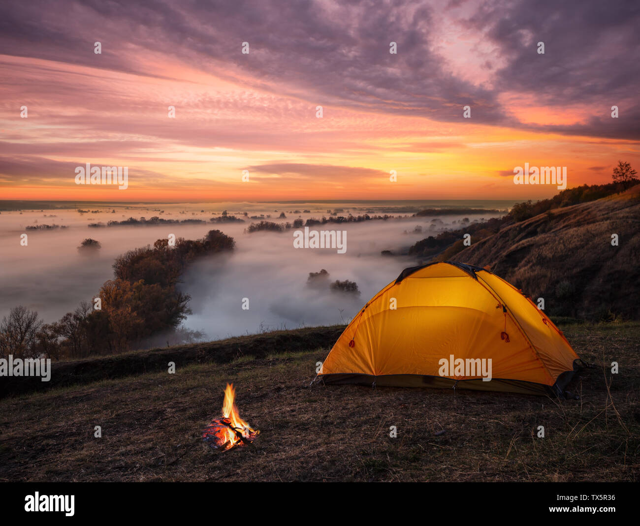 Orange lit inside tent and fire over misty river at sunset Stock Photo