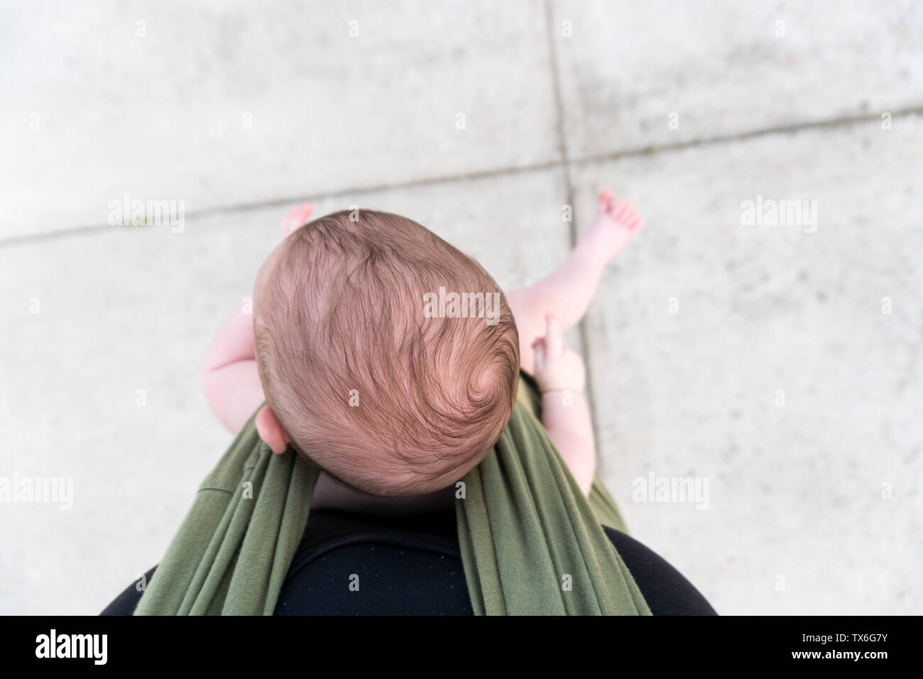 Overhead view of a baby's head with cowlick and foot sticking out as he sits in a green baby carrier of the photographer. Stock Photo