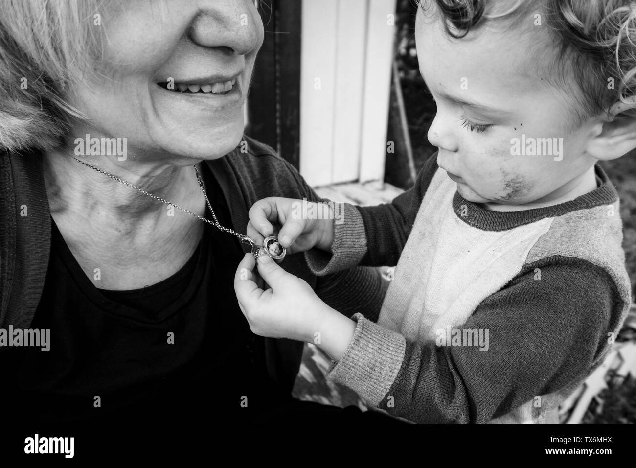 Black and white image of a toddler boy looking at the photo in an open locket hanging around an adult smiling woman's neck. Stock Photo